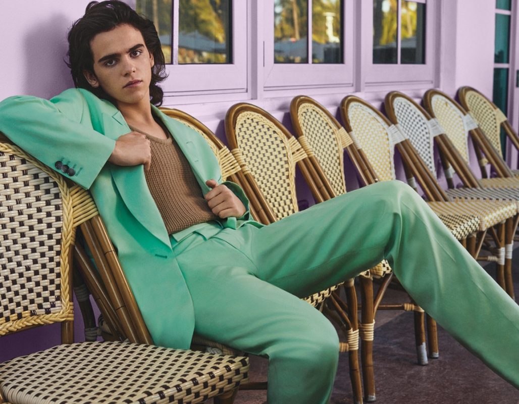 Fashionable young man in a mint green suit poses while seated on a woven chair, set against a pastel purple background with matching window frames.