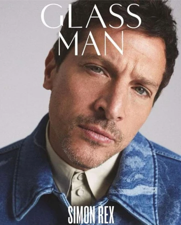 Close-up portrait of actor Simon Rex wearing a blue denim jacket over a collared shirt against a pale grey background.