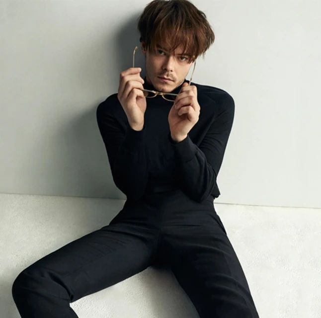 Portrait of a dark-haired adult male in a black turtleneck and pants, holding gold-rimmed glasses, sitting against an off-white wall.