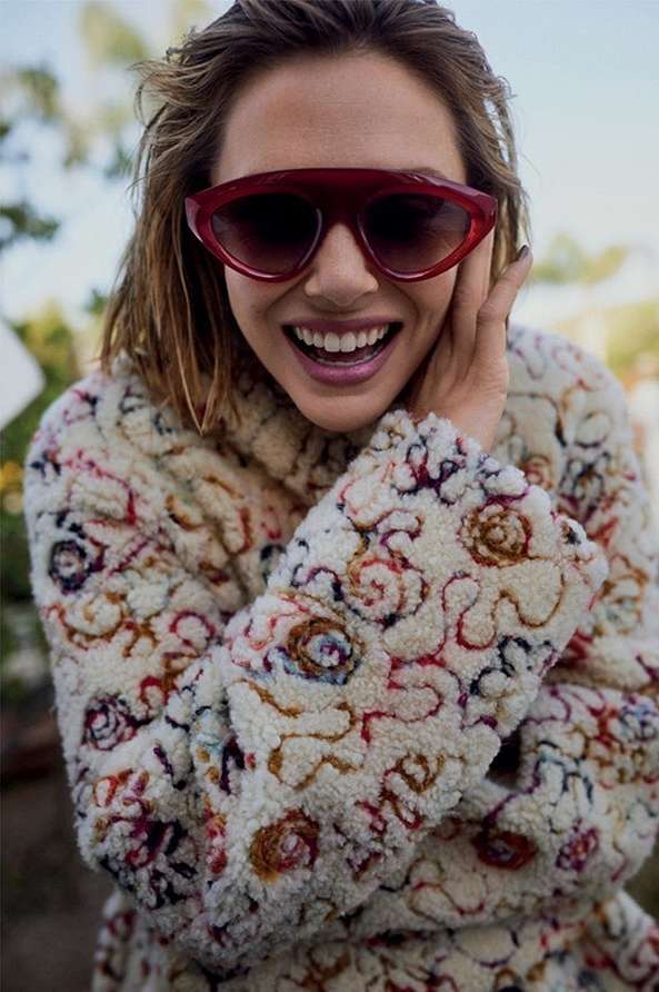 A smiling woman with short hair wears oversized sunglasses and a patterned coat outdoors, hand near her face.