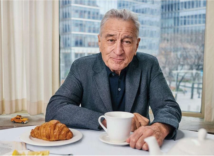 Portrait of elderly adult man with gray hair in grey suit at table with teacup and croissant in front of modern building backdrop.
