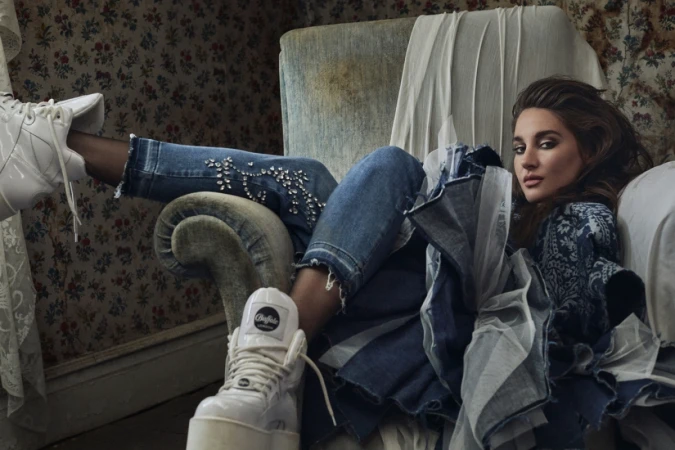 Woman reclining on an armchair wearing embellished denim jeans and platform sneakers against floral wallpaper