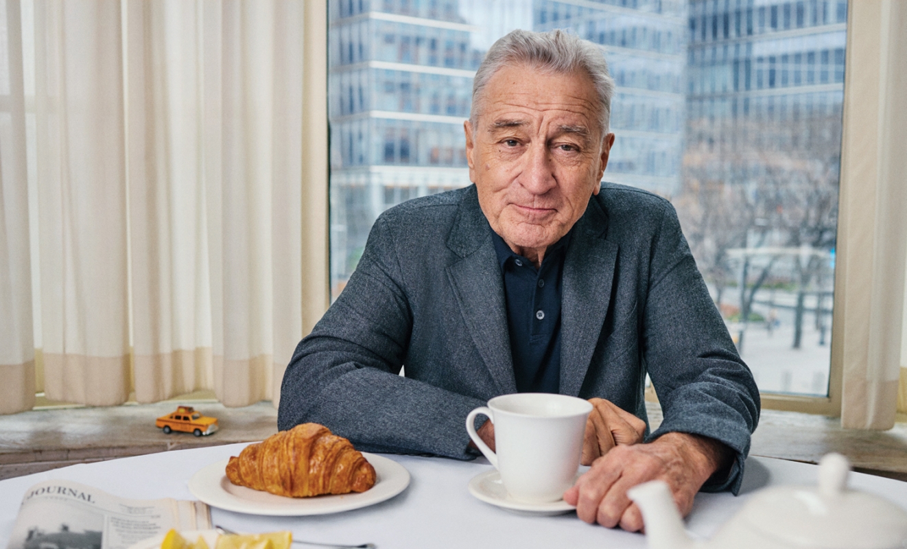 Close-up of an elderly man in a blue blazer sitting at a table with a croissant, cup of coffee, and newspaper in a bright room.