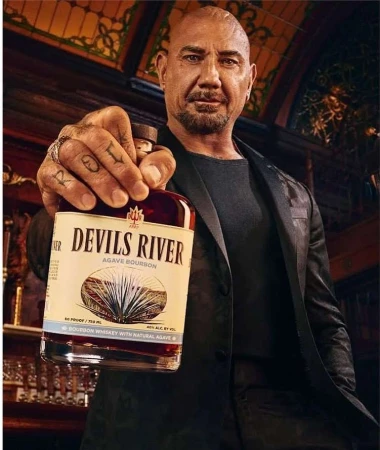 Dave Bautista holds a Devil's River Agave Bourbon bottle in a semi-lit interior setting, showcasing its label, agave art, and design.
