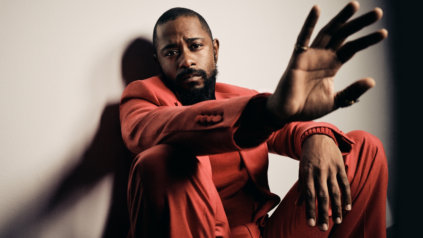 Black man with a beard wearing a red suit seated against a white wall, holding one hand up toward the camera lens.