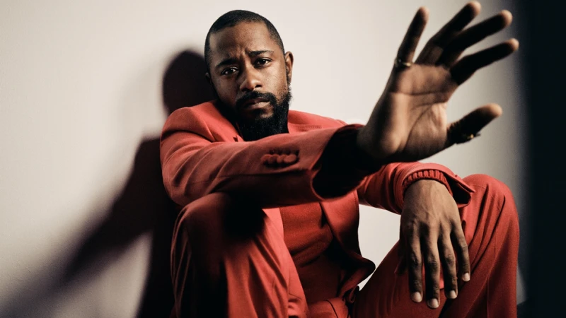 Black man with a beard wearing a red suit seated against a white wall, holding one hand up toward the camera lens.