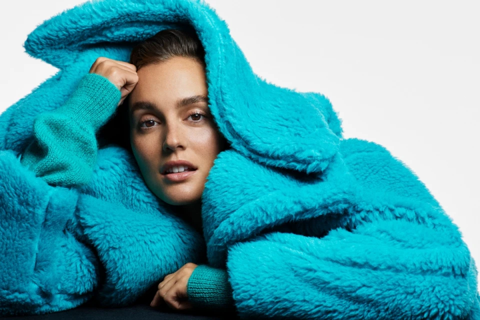 Close-up of a woman with light skin, wearing a fuzzy turquoise coat and sweater, posing against a bright white backdrop.