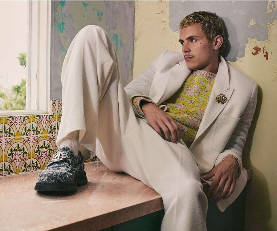 Stylish adult male in a floral sweater and white suit, relaxing on a ledge in a neutral-toned indoor setting with patterned tiles and aged walls.