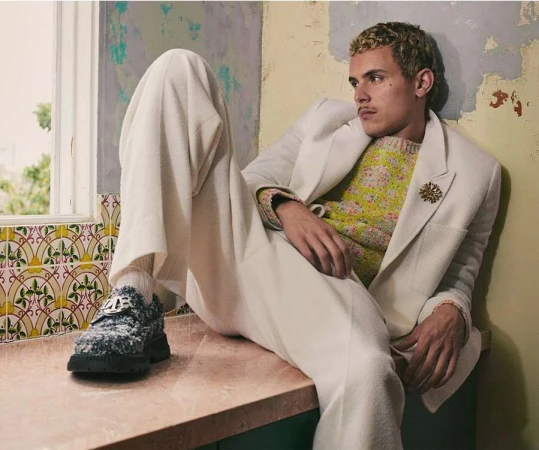 Stylish adult male in a floral sweater and white suit, relaxing on a ledge in a neutral-toned indoor setting with patterned tiles and aged walls.