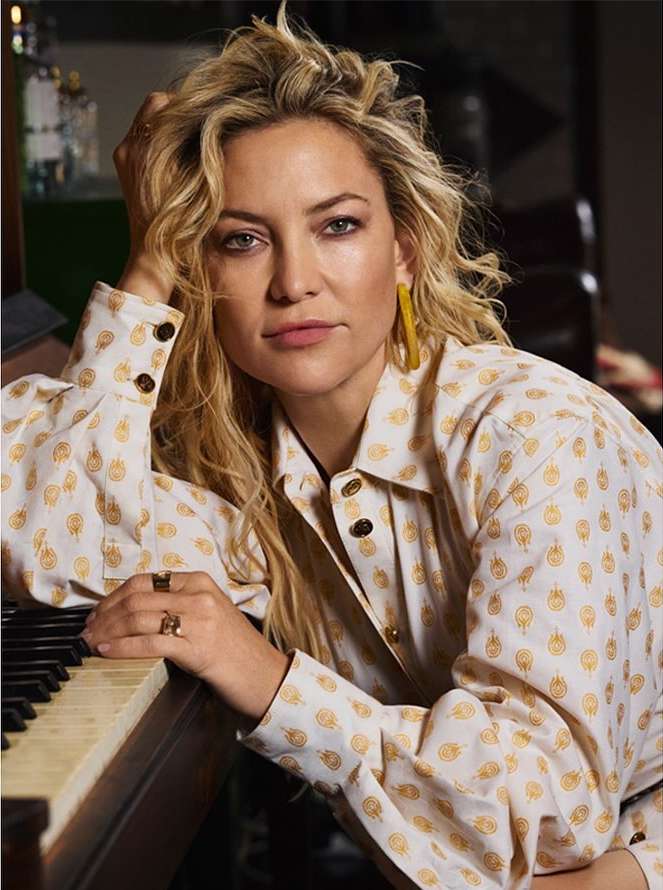 Close-up portrait of a blonde woman wearing a patterned collared shirt and yellow earrings, resting her arm on a piano.