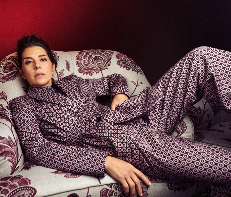Woman with dark hair lies on a patterned settee, wearing a patterned purple suit jacket and matching pants in an indoor setting.