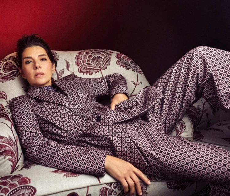 Woman with dark hair lies on a patterned settee, wearing a patterned purple suit jacket and matching pants in an indoor setting.