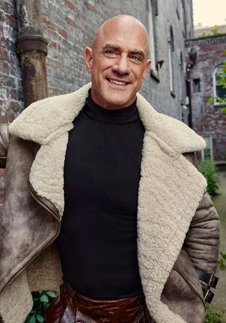 Close-up of bald adult male with a warm smile, wearing a black turtleneck and brown shearling coat in an outdoor urban setting.