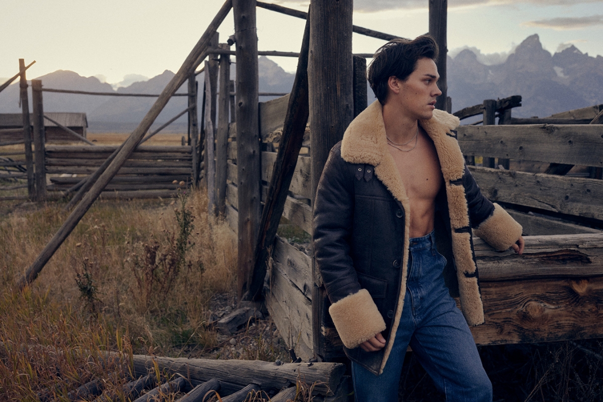 Male model poses in shearling coat against a rustic wooden fence, with mountains in the background on a ranch at golden hour.