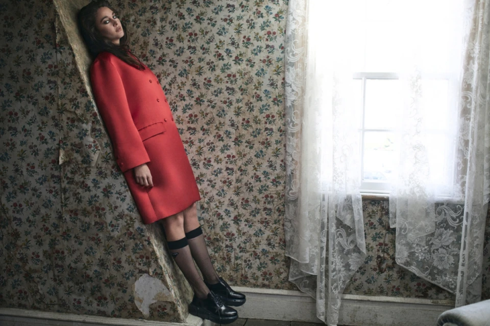 A woman in a red double-breasted coat leans against a floral wallpapered wall in a room with a window covered by sheer lace curtains.