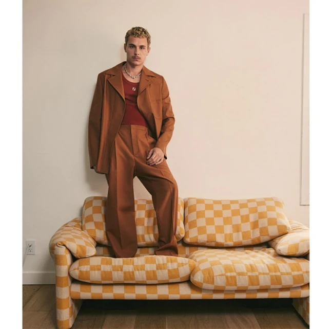 A blonde male model in a brown suit poses on a checkered couch indoors, against a neutral wall for a fashion-forward editorial shot.