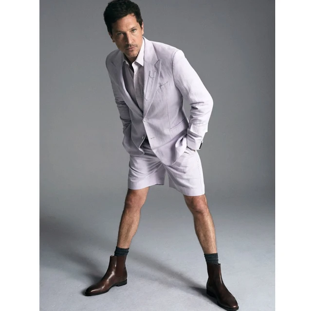 Adult male fashion model poses in a light purple suit jacket and shorts with brown leather boots against a neutral gray backdrop.