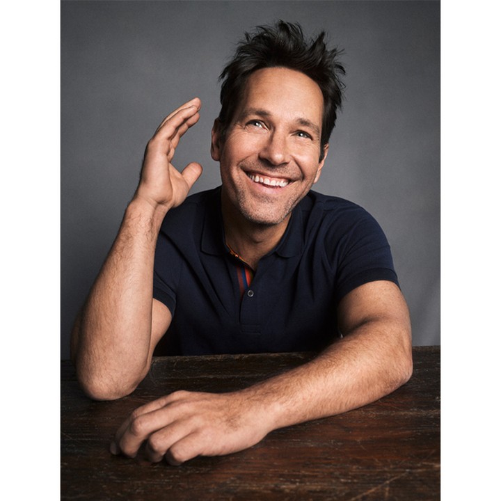 Portrait of actor Paul Rudd smiling and sitting at a wooden table with a grey background. He is wearing a short sleeved polo shirt.