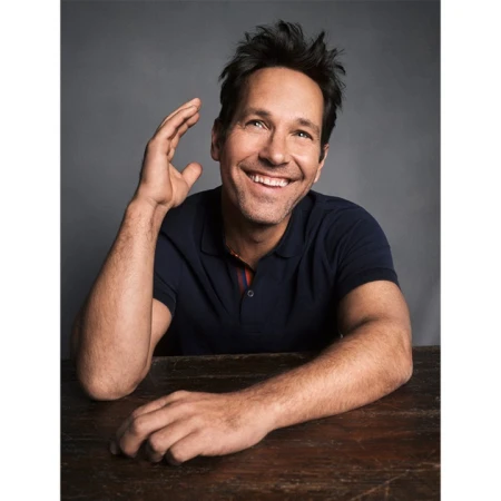 Portrait of actor Paul Rudd smiling and sitting at a wooden table with a grey background. He is wearing a short sleeved polo shirt.