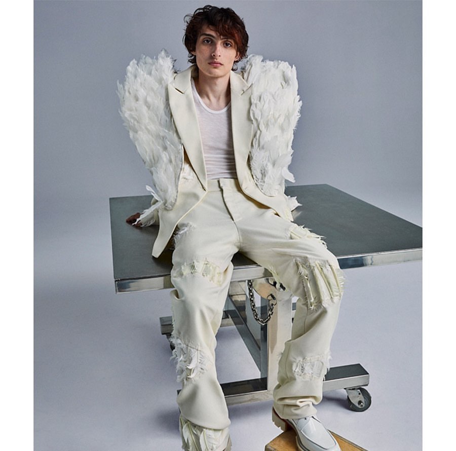 Young man in feather-embellished blazer and distressed pants sits on a steel table in a fashion shot against a solid grey backdrop.