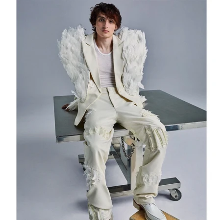 Young man in feather-embellished blazer and distressed pants sits on a steel table in a fashion shot against a solid grey backdrop.