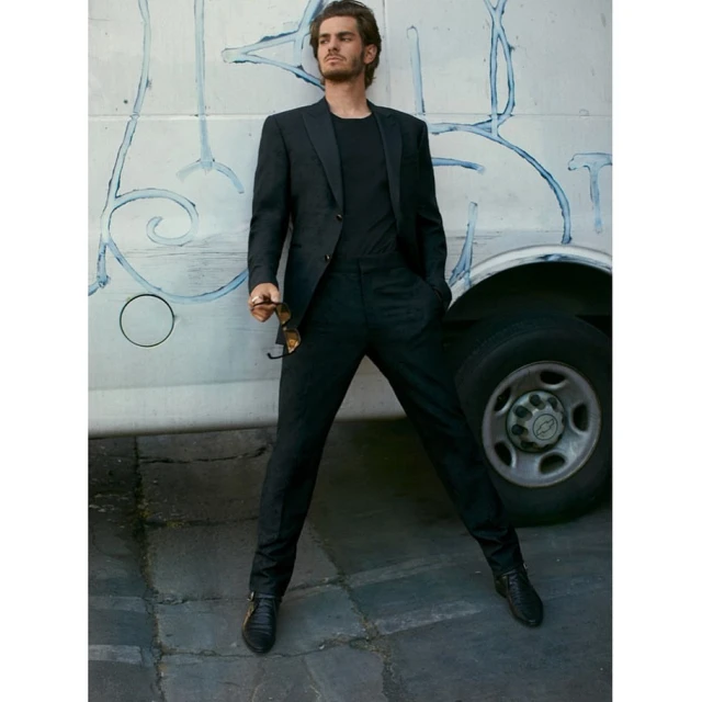 Andrew Garfield poses in a black suit with sunglasses in hand in front of a bus with graffiti, showcasing a blend of sophistication.
