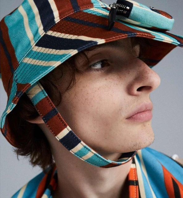 Close-up of a person wearing a striped bucket hat with side straps and a matching striped shirt, posing indoors in soft lighting.