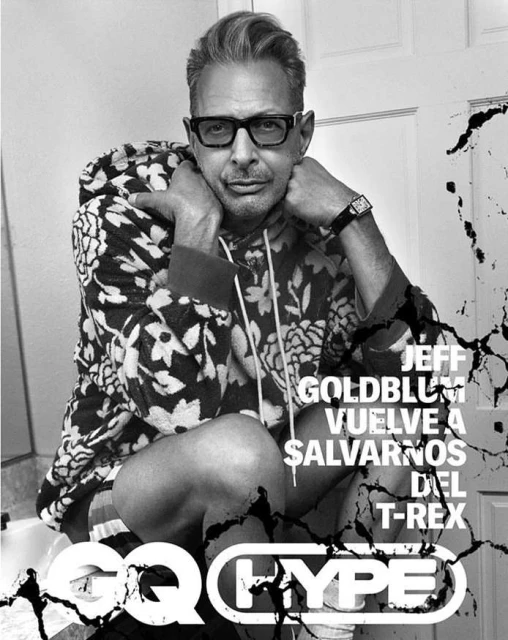 Black and white portrait of Jeff Goldblum in patterned hoodie, glasses, and watch, posing indoors for a magazine cover in a bathroom.
