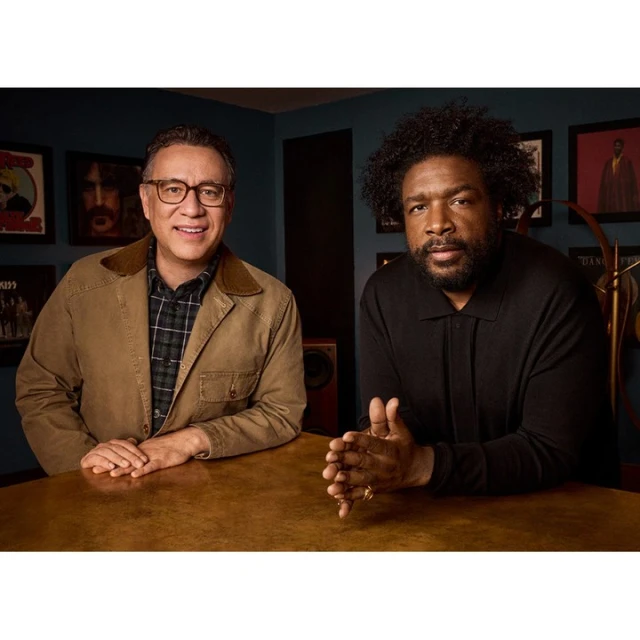 Two adult men sit at a wood table in a room with framed images; one man wears a tan jacket, the other a black shirt. 