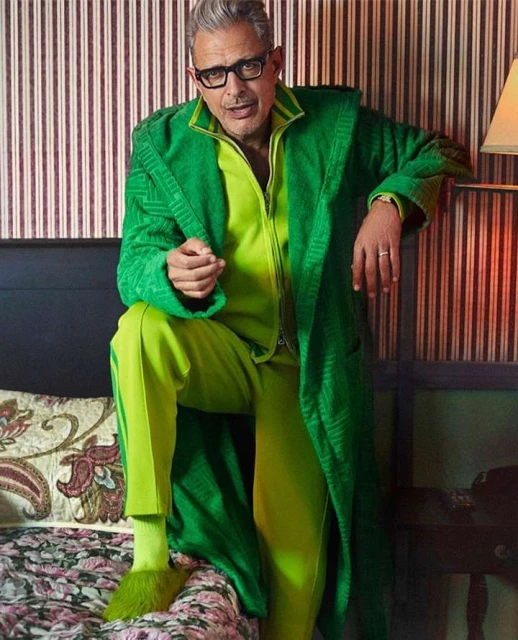 Stylish older man, Jeff Goldblum, wearing a green tracksuit and robe, poses indoors against a striped wall and floral-patterned bed.
