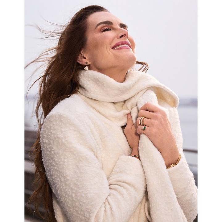Portrait of Brooke Shields smiling with eyes closed, wearing a cream textured coat and holding a matching scarf.