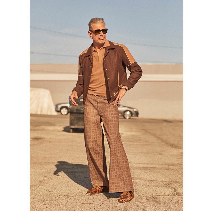 Full-length shot of adult male, Jeff Goldblum, outdoors in brown jacket, patterned pants, and sunglasses with neutral facial expression.