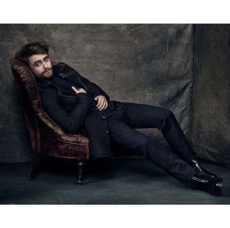 Daniel Radcliffe reclining in a velvet armchair wearing a dark coat and trousers against a textured backdrop.
