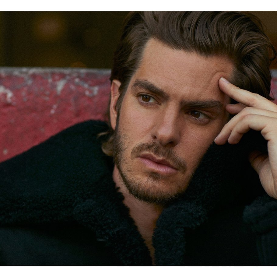 Close-up portrait of Andrew Garfield with stubble wearing a dark textured jacket, touching his temple thoughtfully.