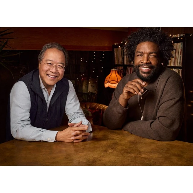 Two adult men, one with an Afro, sit at a brown table indoors. They are smiling, looking directly at the camera, with warm lighting.