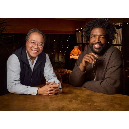 Two adult men, one with an Afro, sit at a brown table indoors. They are smiling, looking directly at the camera, with warm lighting.