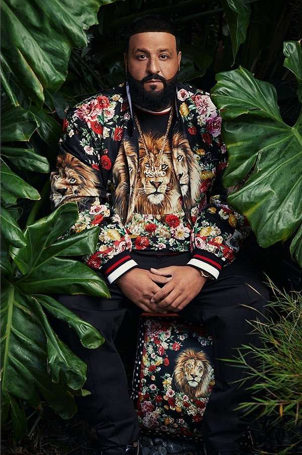 DJ Khaled seated outdoors amidst lush tropical foliage. He wears a lion and floral-themed jacket and pants. Close-up, eye-level shot.
