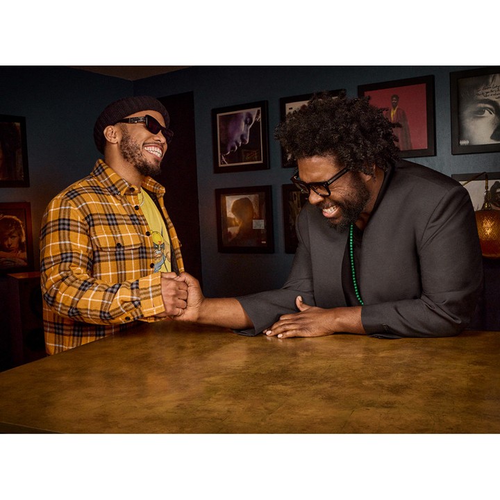 Two adult males indoors, shaking hands at a table. One wears a plaid shirt and hat, the other a suit. Framed art hangs on the wall.