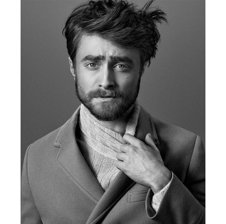 Portrait of Daniel Radcliffe with beard and tousled hair wearing a coat and scarf against a grey background