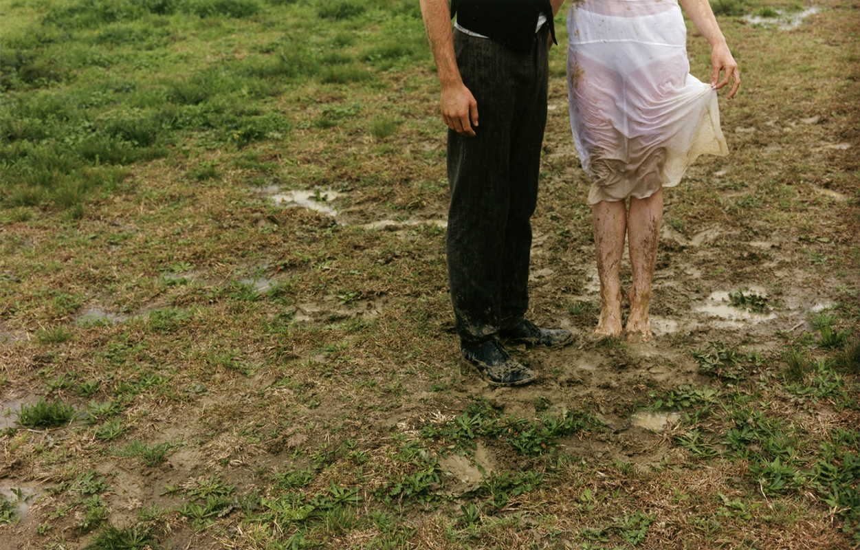 A couple stands barefoot in a muddy field, the woman lifting her white dress, capturing a raw and natural moment outdoors.