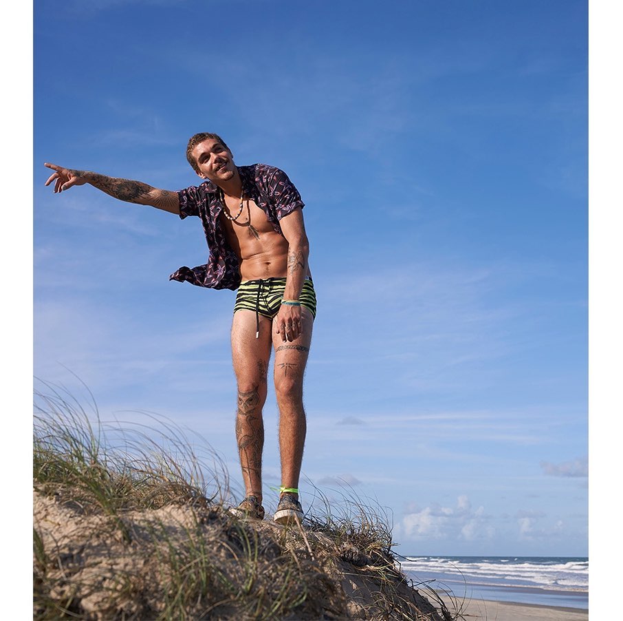 Athletic adult male with tattoos stands on a sandy hill with open button-down shirt, swimwear, pointing towards the ocean under a blue sky.