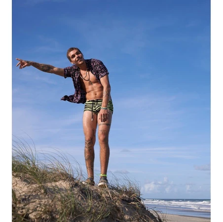 Athletic adult male with tattoos stands on a sandy hill with open button-down shirt, swimwear, pointing towards the ocean under a blue sky.