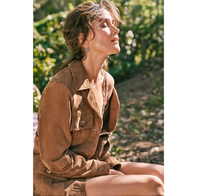 Side view of woman in brown suede jacket and shorts sitting outside with eyes closed enjoying sunshine in a natural outdoor setting.