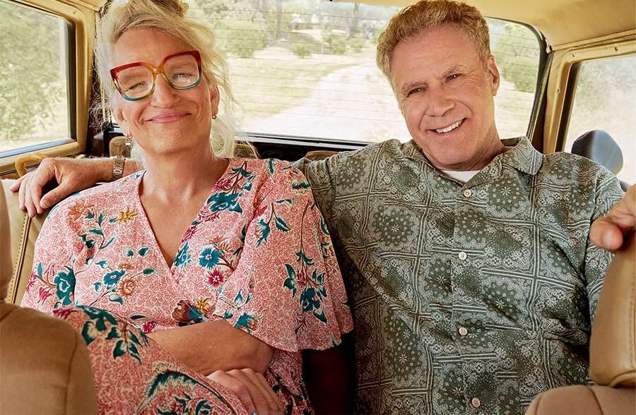 Indoor shot of a smiling adult woman with colorful glasses and a patterned dress sitting next to a smiling adult man in a patterned shirt.