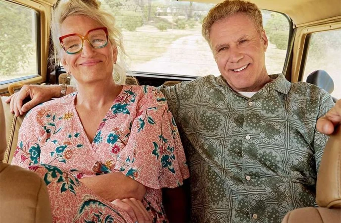 Indoor shot of a smiling adult woman with colorful glasses and a patterned dress sitting next to a smiling adult man in a patterned shirt.