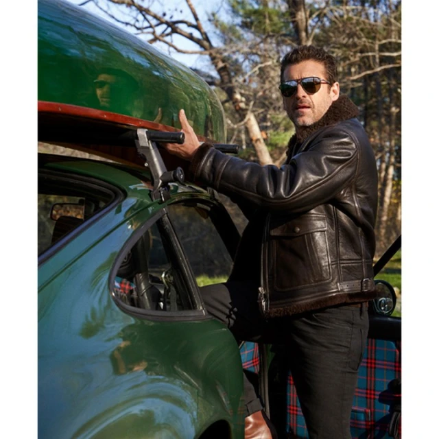 Adult man in leather jacket loads green car with canoe on roof rack; outdoor setting with trees in background on a sunny day.