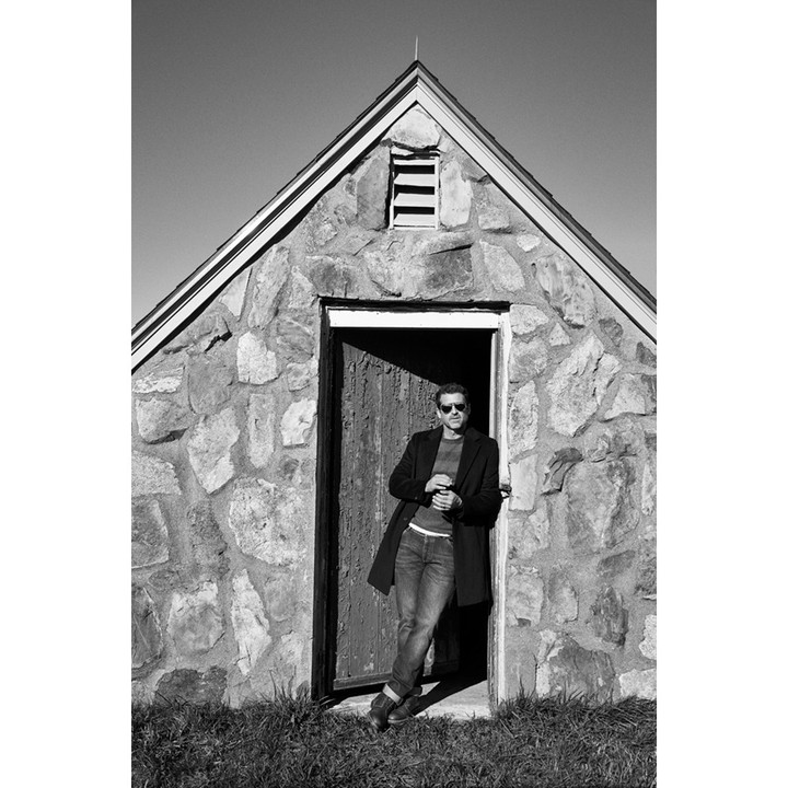 Monochrome portrait of a man in sunglasses leaning against a stone structure, wearing a dark coat and jeans, on a sunny day.