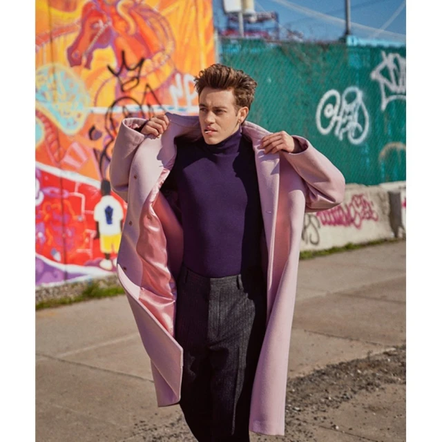 An adult man in a purple turtleneck and grey pants models a light pink overcoat on a city street in front of a graffiti wall.
