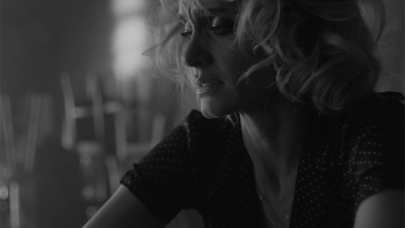 Black and white close-up portrait of a blonde woman with wavy hair wearing a patterned top, captured by Carter Smith.