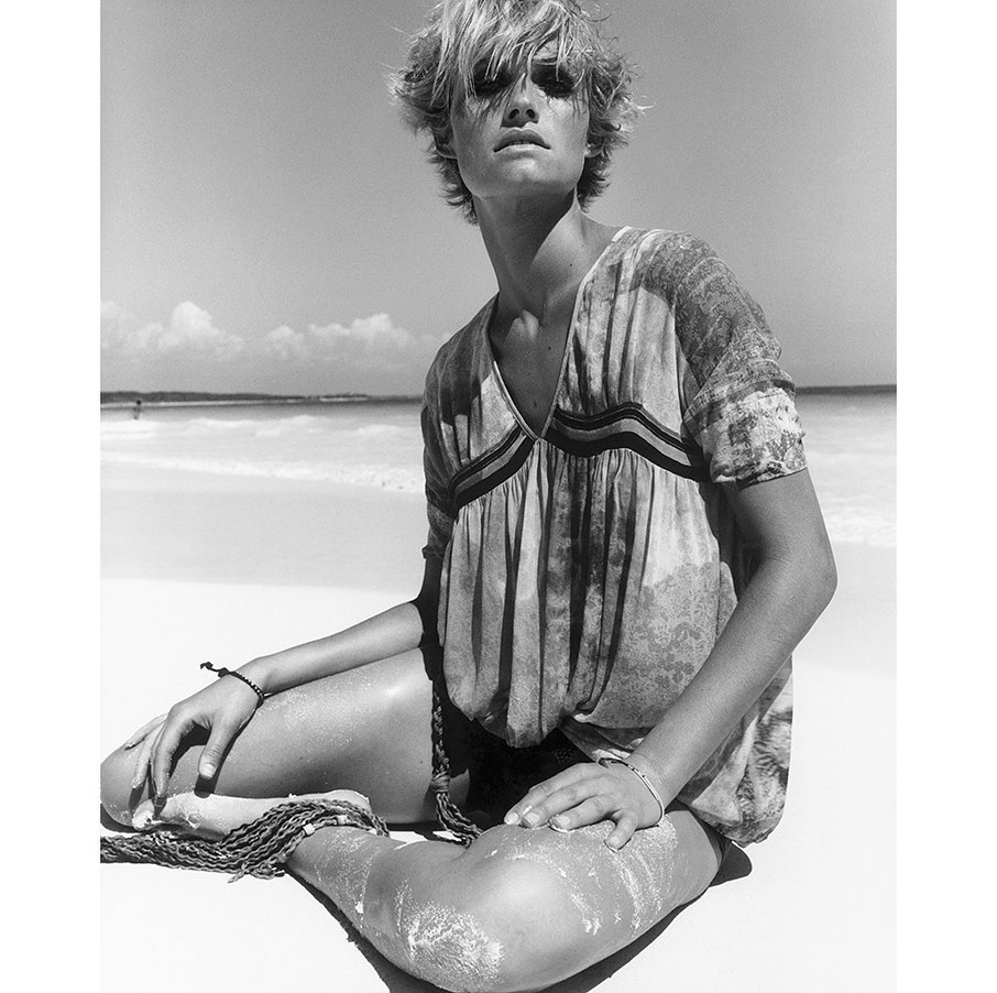 Black and white portrait of a blonde model with short tousled hair sitting cross-legged on a sandy beach